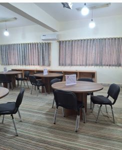Bright interior of a dining/meeting room with round wood tables, black chairs, striped carpet, and large curtained windows; ceiling lights and an wall-mounted air conditioner visible.