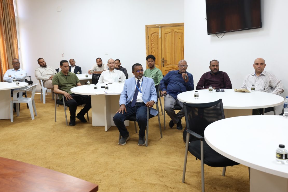 Group of men seated around white round tables in a meeting room, facing the camera.