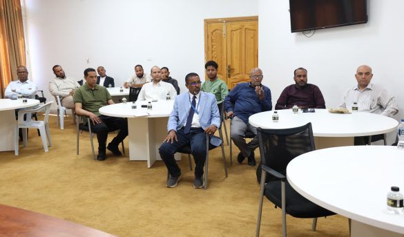 Group of men seated around white round tables in a meeting room, facing the camera.