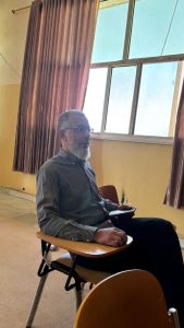 Middle-aged man with glasses and gray beard sitting in a classroom chair with a folding desk, taking notes.