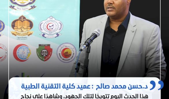 Man in a light gray suit speaks at a podium with a microphone, a banner with logos behind him, and a white speech-bubble with Arabic text at the bottom.