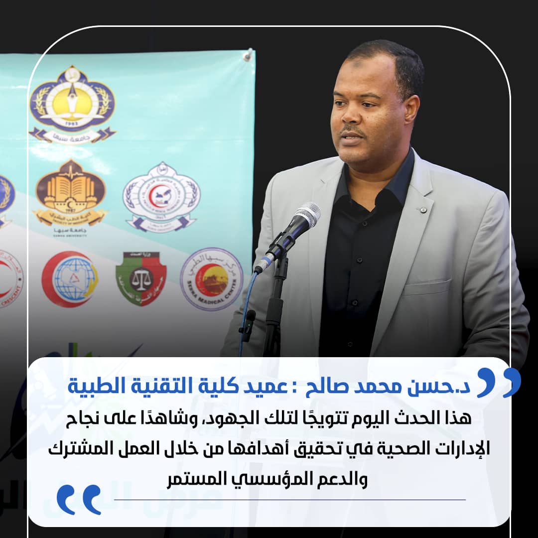 Man in a light gray suit speaks at a podium with a microphone, a banner with logos behind him, and a white speech-bubble with Arabic text at the bottom.