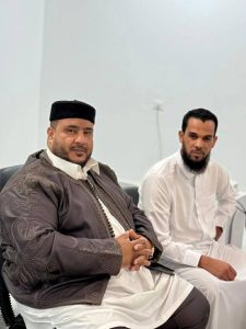 85126502-ad19-463b-b42c-74ab169406fd - جامعة سبها | Sebha University Two men in traditional attire sit indoors; left man wears an embroidered brown robe and black cap, right man wears a white thobe.
