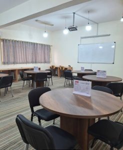 Round wooden tables with black chairs scattered in a training room; whiteboard on the wall and a ceiling projector above.