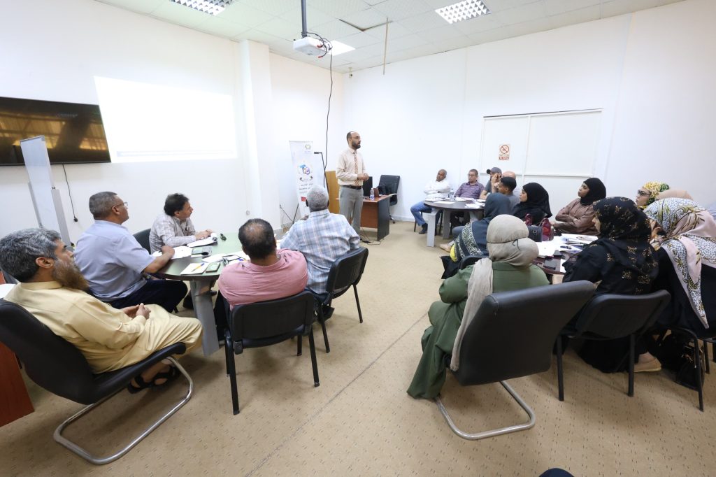 Man presenting at the front of a classroom to a diverse group of adults seated around tables; projector screen displayed on the left side of the room.