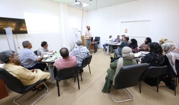 Man presenting at the front of a classroom to a diverse group of adults seated around tables; projector screen displayed on the left side of the room.
