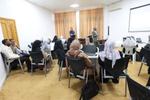 Workshop scene: attendees in hijabs seated around round tables with laptops as two presenters speak at the front near a projection screen.