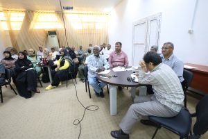 Diverse group of people seated in a conference room, listening to a presentation around a table with papers and notebooks.