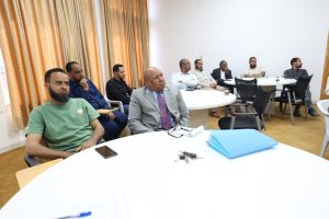 Several men sit around white round tables in a meeting room, listening attentively during a discussion or presentation.