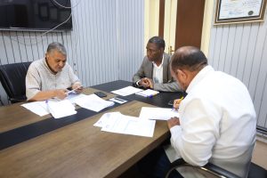 Three men seated around a conference table reviewing documents, with papers, notebooks, and phones on the table in a small office setting.