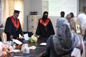 Graduates in black gowns and red sashes stand at a table during a formal graduation ceremony, with attendees watching a projection on the wall.