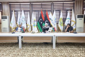 Three officials seated at a conference table with flags and emblems behind them, during a formal meeting.
