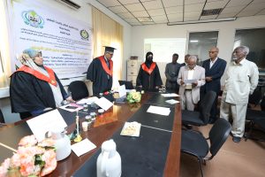 Group of academics in graduation gowns and red stoles stand around a conference table during a formal ceremony in a classroom or meeting room. A large banner with Arabic text and logos hangs on the wall behind them, indicating an academic event or workshop.