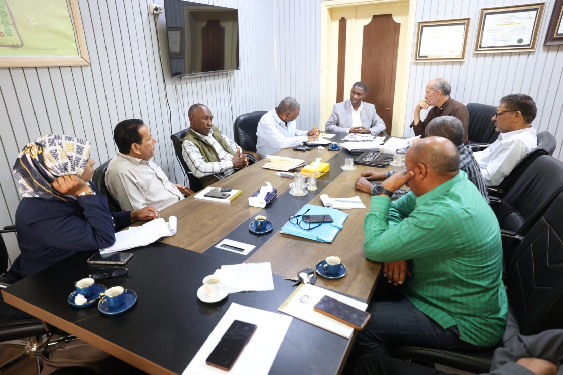 Group of professionals seated around a long conference table, engaged in a meeting with papers and notebooks nearby.