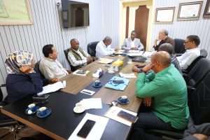 Group of professionals seated around a long conference table, engaged in a meeting with papers and notebooks nearby.