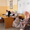 Five adults sit at a wooden classroom desk, women in hijabs and a man using a tablet, with a whiteboard and notes in the background.