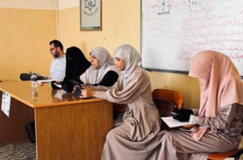 Five adults sit at a wooden classroom desk, women in hijabs and a man using a tablet, with a whiteboard and notes in the background.