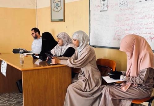 Five adults sit at a wooden classroom desk, women in hijabs and a man using a tablet, with a whiteboard and notes in the background.
