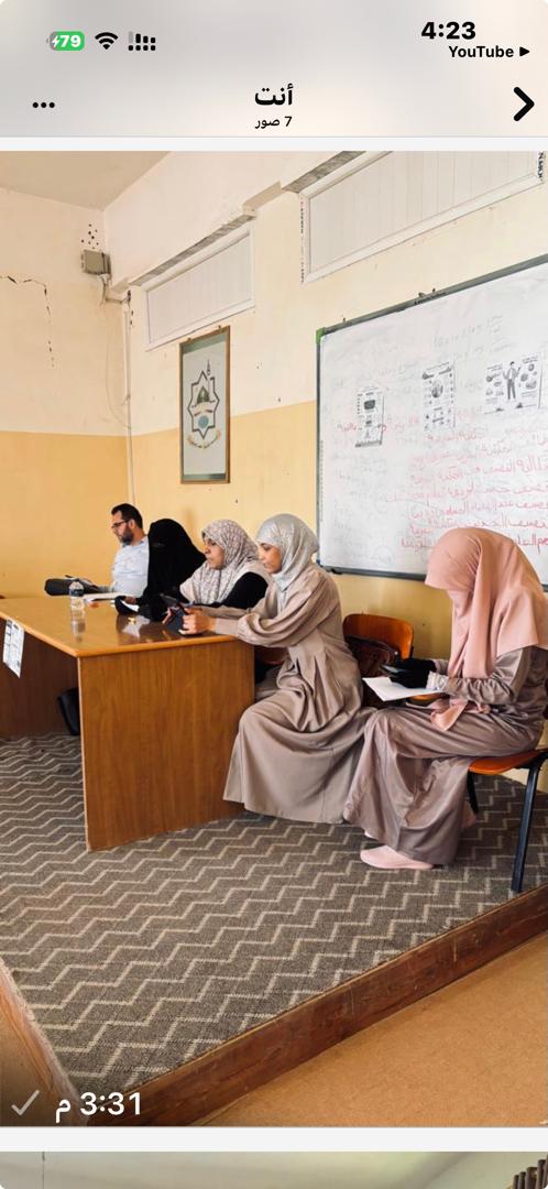 Five adults sit at a wooden classroom desk, women in hijabs and a man using a tablet, with a whiteboard and notes in the background.