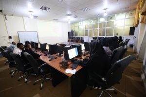 Group of students wearing hijabs sit around a U-shaped desk with desktop computers in a computer lab, a whiteboard and projector at the front.