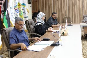 Group of professionals seated at a long conference table with microphones and flags in the background.