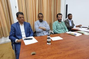 Four men in business attire sit at a wooden conference table, engaged in a meeting with papers, pens, and bottles on the table behind beige curtains.”] ,