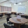 Circular wooden tables with black chairs in a classroom or meeting room, whiteboard on wall, projector lights overhead, striped carpet.