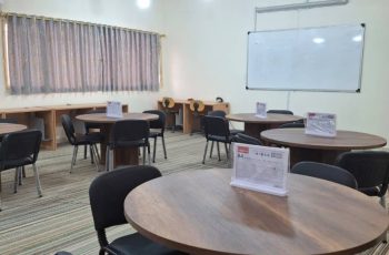 Circular wooden tables with black chairs in a classroom or meeting room, whiteboard on wall, projector lights overhead, striped carpet.