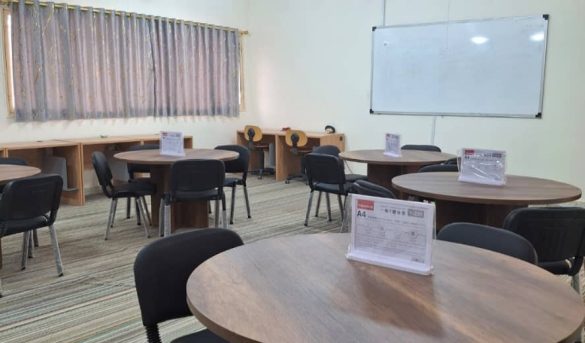 Circular wooden tables with black chairs in a classroom or meeting room, whiteboard on wall, projector lights overhead, striped carpet.