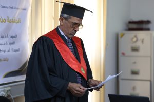 Man in a black graduation gown and cap reads a document at a ceremony, with a banner in Arabic in the background.