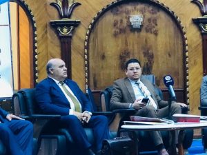 Panel discussion with three men in suits seated on a stage, microphones and documents on a table in front of them, formal conference setting.