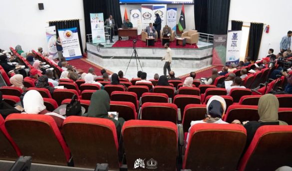 Panel discussion on stage with banners and flags, audience seated in red theater chairs in a conference hall.