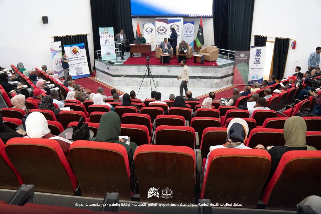 Panel discussion on stage with banners and flags, audience seated in red theater chairs in a conference hall.