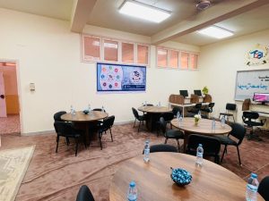 Conference room with round tables, black chairs, water bottles, and a poster on the wall; laptops on a back desk and a plant in the corner.