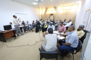 Male speaker presenting to a diverse group of adults in a training room with beige curtains in the background.