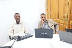 Two men sit at a white table with laptops in a meeting; one wears a beige shirt, the other a tan blazer, with a wooden door in the background.