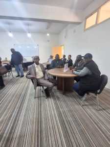 Group of men seated around a round wooden table in a bright conference room with a whiteboard in the background.