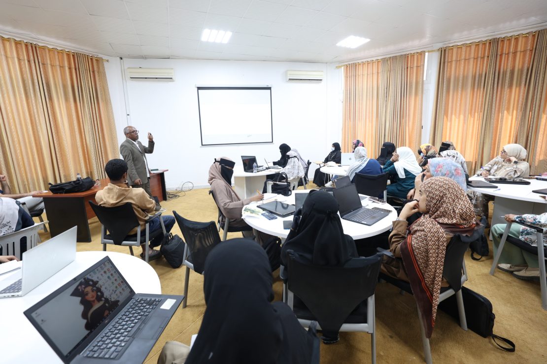 A male presenter stands at the front of a classroom, gesturing while addressing a group of people seated around round tables with laptops and notebooks.