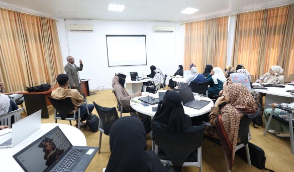 D9124120-954D-49AC-B977-CAB3339E75B8 - جامعة سبها | Sebha University A male presenter stands at the front of a classroom, gesturing while addressing a group of people seated around round tables with laptops and notebooks.