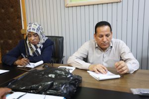 Two adults sit at a conference table signing papers; woman in a patterned hijab on the left, man in a light shirt on the right.