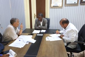 Three men seated around a conference table in a meeting room, discussing documents on the table.