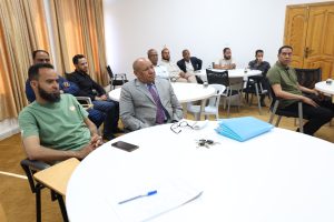 Group of men seated around white round tables in a meeting room, listening attentively; papers, folders, a notebook and keys on the foreground table.