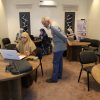 A training room with women in hijabs working on laptops around round tables; a man stands by a table, supervising or guiding them.
