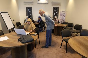 A training room with women in hijabs working on laptops around round tables; a man stands by a table, supervising or guiding them.