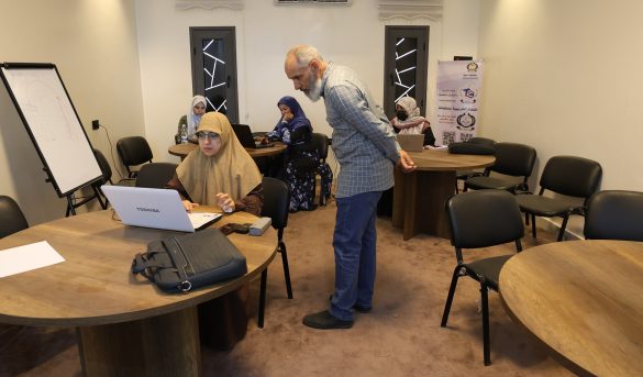 A training room with women in hijabs working on laptops around round tables; a man stands by a table, supervising or guiding them.