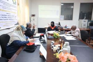 Five professionals around a conference table in a meeting room; projector shows slides and a banner with Arabic text on the wall.
