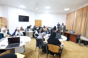 A classroom training session with attendees at round tables using laptops while a presenter speaks at the front of the room.