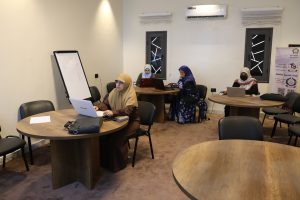 F05158B1-BE84-476E-A6DF-FA7E04AD9147 - جامعة سبها | Sebha University Three women in hijabs work on laptops around round wooden tables in a meeting room; a whiteboard stands to the left.