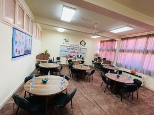 Training room set up for a workshop: round tables with bottled water and black chairs, banners and logos on the back wall, pink curtains.