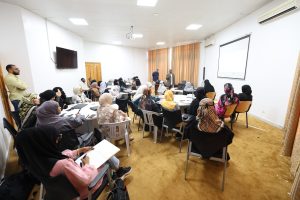 Conference room full of attendees, mostly women in hijabs, listening to two presenters at the front with a projector screen overhead.